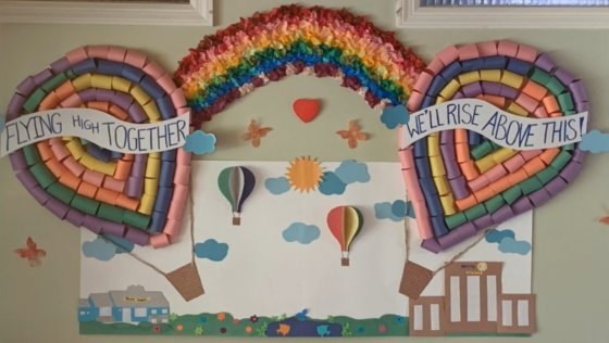 Balloons and Rainbow decoration on bulletin board that says Flying High Together We will Rise above this|Residents showing excitement|Photo of man in yellow plaid shirt||Cares Act with Amer|adult residents coloring around a table|man holding flowers and balloons standing in doorway|staff in PPE standing in hallway of the recovery hub